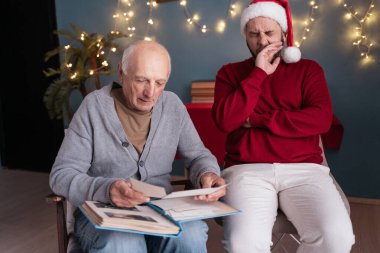 Yaşlı babası yılbaşı fotoğraflarıyla fotoğraf albümüne bakarken Noel Baba şapkalı sıkılmış adam sandalyeye oturmuş esniyor. Boşluğu kopyala