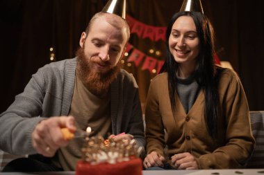 Parti şapkalı genç bir çift masanın yanında oturuyor ve doğum günü pastasında mum yakıyor, evde birlikte kutluyorlar. Aile yıl dönümü partisinde eğleniyor. Boşluğu kopyala