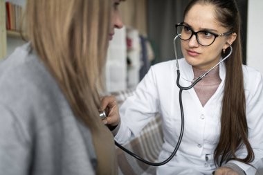 Kalp ve akciğer kontrolü. Steteskop kullanan bayan doktor yaşlı bir kadının nefesini ve evdeki koltukta oturan kalp atışlarını dinliyor. Boşluğu kopyala