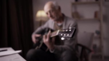 Yaşlı müzisyen gitarda teller kullanıyor. Emekli erkek evde gitar çalıp pratik yapıyor.