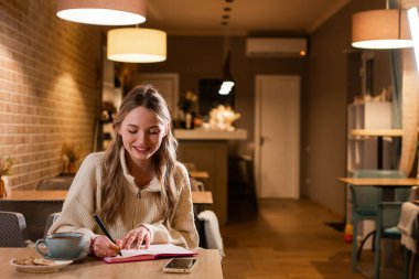 Sarışın, güzel bir kadın, kendi günlüğüne bir fincan kahveyle rahat bir kafede dinlenip, not defterine iyi bir fikir ekliyor. Boşluğu kopyala