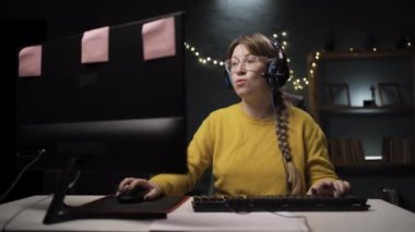 Gözlüklü kadın oyuncu kişisel bilgisayarda online video oyununda oynuyor. Profesyonel oyuncu şampiyonluktan zevk alıyor. Kafasını kaşıyor. Kollarını uzatıyor.