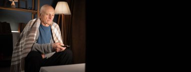 Gri saçlı yaşlı bir adam akşamleyin oturma odasındaki kanepede dinleniyor televizyon izliyor ve uzaktan kumandayla kanal değiştiriyor.