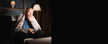 Geceleri elinde uzaktan kumandayla televizyon izleyen yaşlı bir adam ciddi bir ifadeyle düşünceli bir şekilde ekrana bakıyor..