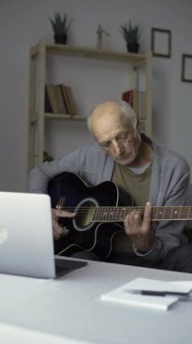 Emeklilik hobileri ve boş vakitleri. Çevrimiçi öğrenme dersleri. Yaşlı bir adam gitar çalma üzerine bir video dersi izler, evdeki bir sandalyeye oturur. Boşluğu kopyala.
