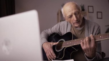 Emeklilik hobileri ve boş vakitleri. Uzaktan öğrenme ve dersler. Yaşlı adam gitar çalma üzerine video dersi izliyor, online öğretmenle sandalye danışmanlığı yapıyor, parmaklarını tellere koyuyor..