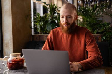 Kafedeki masada oturan odaklanmış kızıl saçlı sakallı adam milenyumu. ciddi bir işadamı bilgisayarla uzaktan çalışıyor veya çalışıyor, klavyede daktilo kullanıyor.