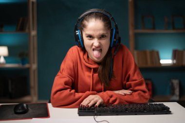 E-sport female gamer play video game at home using pc. Brunette woman gaming player showing tongue feeling negative disgust emotion, broadcast live streaming online in the living room. Webcam view