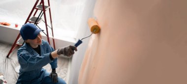 Bir sanatçı boya silindiriyle duvar boyar. İnşaat işçisi yüzeyi açık renklerle boyuyor.