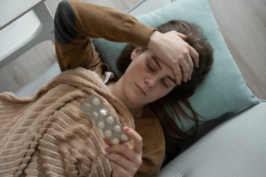 Soğuk algınlığı, grip ve enfeksiyon konsepti. Kareli kumaş giyen genç bir kadın oturma odasındaki kanepede yatarken baş ağrısı ve ateş çekiyor. Üst görünüm