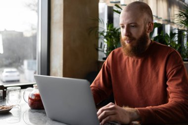 Kafedeki masada oturan odaklanmış kızıl saçlı sakallı adam milenyumu. ciddi bir işadamı bilgisayarla uzaktan çalışıyor veya çalışıyor, klavyede daktilo kullanıyor.