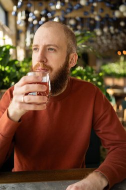 Sakallı, turuncu kazaklı, kızıl saçlı adam pencereden dikkatlice bakarken çay içer. Sıcak atmosferde çay saatlerini sever, bir anlık sakinliği ve rahatlığı yansıtır..