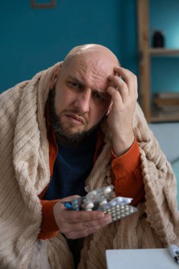 Grip olmuş kel bir adam, sıcak bir battaniyeye sarılmış, elinde ilaç paketleri var. Düşünceli bir ifade, baş ağrısı ve ateşten kurtulmak için. Konforlu ev ortamı iyileşme çabalarını yansıtıyor.