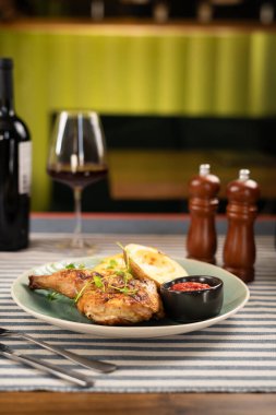 Kızarmış patates yarımları ve yanında baharatlı kırmızı sos ile servis edilen sulu ızgara tavuk bacağı. Yanında da bir kadeh kırmızı şarap. Restoran ortamında mükemmel bir yemek deneyimi yaratıyor..