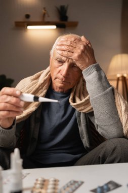 Sıcak eşarp takan, termometreyi tutan, sıcaklığı ölçen, alnına dokunan, evde kendini kötü hisseden, ilaçlarla çevrili, sıcak akşam ışığı ile aydınlanan yaşlı bir adam..