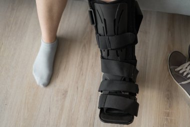 Siyah ortopedik bot giyen yaralı bir bacağın yakın çekimi, iyileşme ve rehabilitasyonu vurguluyor. Diğer ayağının çorabın içinde olması ve ayakkabının yanında olması ev ortamını işaret ediyor..