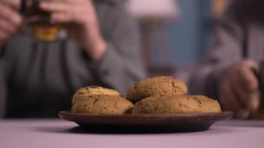 Ön plandaki ahşap tabakta kurabiyelerin yakın çekimi. Bulanık arka planda çay içip kurabiye yiyen iki insan rahat ve sıcak bir ortam yaratıyor..