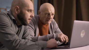 Yetişkin oğlu emekli babasına sosyal medya, online alışveriş ve ödemeler için dizüstü bilgisayar kullanmayı öğrenmesine yardım ediyor. İç açıcı bir bağ kurma ve evdeki dijital becerileri paylaşma anı.