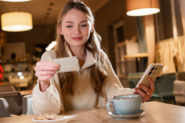 Молодая женщина сидит в уютном кафе в течение вечера, держа в руках кредитку и смартфон. Она сосредоточена на осуществлении онлайн-платежей или покупок в окружении теплой атмосферы.