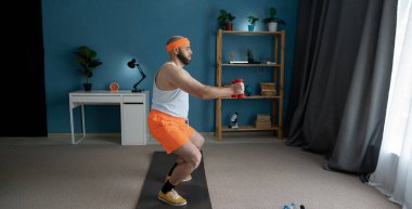 Turuncu şortlu komik bir sporcu spor salonunda dambıllarla çömelme yapıyor, aktif spor yaşam tarzı kavramı var.