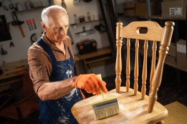Önlük giymiş usta bir zanaatkar ve eldivenler iyi donanımlı atölyedeki fırçayı kullanarak ahşap sandalyeyi dikkatlice boyuyor. Mobilyaları ustalıkla ve ustalıkla restore etmeye adanmıştır..