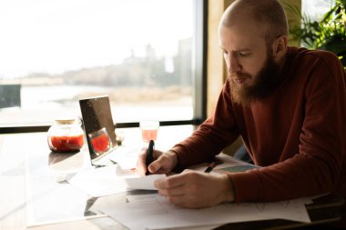 Sakallı işadamı, kafede çalışan dizüstü bilgisayarını kullanarak çalışan kağıt belgeleri not alıyor. Boşluğu kopyala