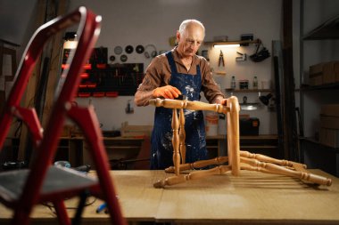 Yaşlı erkek marangoz, kumlu bacakları zımparalıyor. Koyu marangozluk atölyesinde zımpara süngeri yenileniyor. Mobilya restorasyonu, DIY ve hobi konsepti