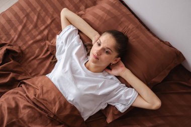 Beyaz gömlekli kadın, kahverengi çarşaflı, elleri başının arkasında uzanmış, düşünceli ve huzurlu görünüyor. Konforlu bir evde rahatlama, rahatlama ve kişisel zaman kavramı.