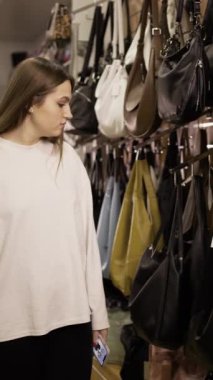 şık butikte el çantalarını karıştıran, yeni koleksiyonu inceleyen, vitrin boyunca yürüyen, akıllı telefonla fiyat etiketini fotoğraflayan, moda mağazasında alışveriş kararı veren bir kadın. Dikey video