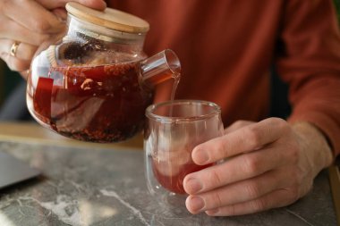Elleri cam çaydanlığı tutan, şeffaf bardağa sıcak meyve çayı dolduran sıcak ve rahatlatıcı bir kafede bir masada oturan bir adamın yakın çekimi..