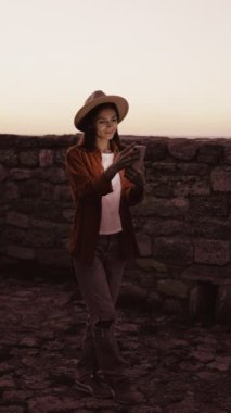 Şapkalı bir kadın, gün batımında taş duvarın karşısında duran cep telefonu ekranı kullanarak sosyal medya okuyor. Boşluğu kopyala