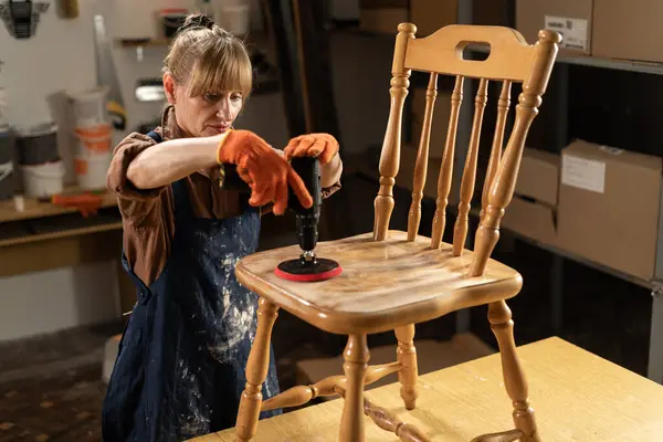 Kadın marangoz, akşamları atölyesinde çalışan elektrikli zımpara makinesiyle ahşap sandalyeyi zımparalıyor. Boşluğu kopyala