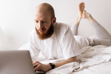 Genç adam dizüstü bilgisayar kullanıyor ve yatak odasında çalışıyor evde yatakta oturuyor. Boşluğu kopyala