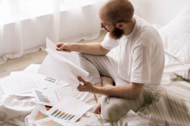 Muhasebe konsepti. Sakallı adam yatak odasının içinde kağıt belgelerle evde çalışıyor. Boşluğu kopyala