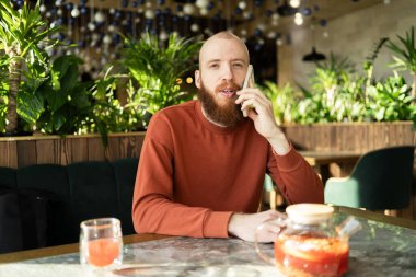 Kızıl sakallı genç bir adam çay molasında kafede otururken cep telefonuyla konuşuyor. Boşluğu kopyala