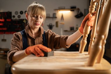 Mobilya yenileme ve diy konsepti. Orta yaşlı bir kadın kumlu süngerle eski ahşap sandalyeyi zımparalıyor. Boşluğu kopyala