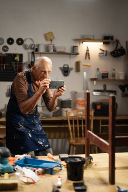 Önlük giyen üst düzey marangoz atölyede akıllı telefonlu mobilya projesinin fotoğrafını çekiyor, restorasyon sürecini belgeliyor, dijital araç gereç kavramı, işçilik ve küçük işletme kavramı