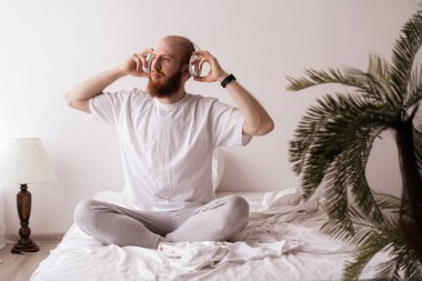 Genç yakışıklı adam beyaz pijama giyiyor ve kulaklık takıyor yatakta müzik dinliyor yatak odasında dinleniyor uyanıyor ve keyifli bir gün geçiriyor. Boşluğu kopyala