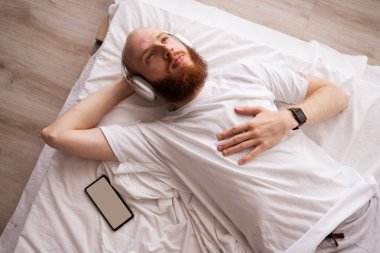 Kulaklık takan, cep telefonlu bir adam sabah radyoyu dinlerken yatakta dinleniyor. Teknoloji, sağlık ve müzik konsepti