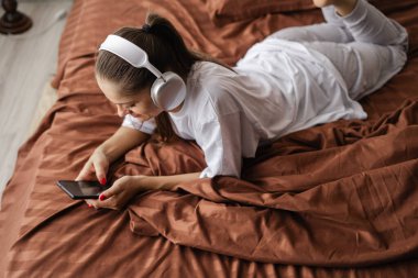 Beyaz kadın akıllı telefon kullanıyor kulaklık takıyor ve yatağında müzik dinliyor. Bayan rahatlatıcı harika bir hafta sonu geçiriyor. Cihazlar ve internet kavramı. Üst görünüm