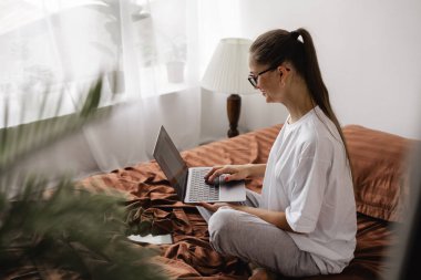 Mutluluktan gülümseyen bir kadın yatak odasında dizüstü bilgisayar kullanarak çalışıyor. Boşluğu kopyala