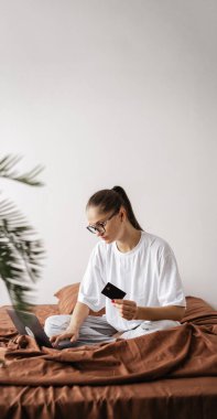 Kredi, ödeme, alım ve alışveriş konsepti. Laptop ve banka kartı kullanan bir kadın yatak odasında online işlem yapıyor. Pankart. Dikey. Boşluğu kopyala