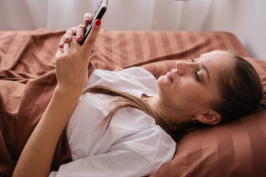 Teknoloji ve insan kavramı. Genç bir kadın yatakta akıllı telefondan mesajlaşarak sohbet ediyor. Sabahları yatak odasında dinleniyor. Yakın plan.