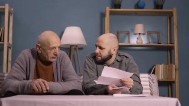 Emekli baba ve yetişkin oğlu evlerinin oturma odasında otururken faturaları okuyorlar. Boşluğu kopyala