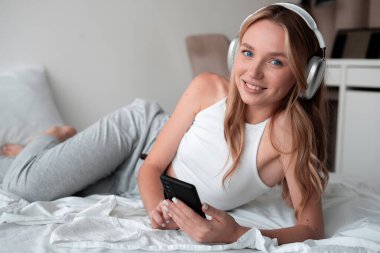 Kulaklıklı genç bir kadın yatak odasında oturmuş müzik dinliyor. Sabahın tadını çıkarıyor. Boşluğu kopyala