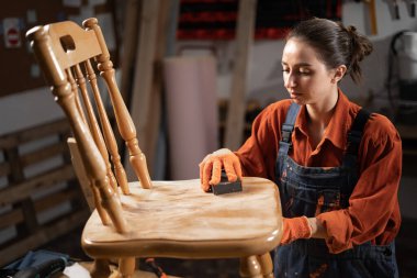 Mobilya yenileme ve diy konsepti. Marangozluk atölyesinde eski ahşap sandalyeyi süngerle zımparalayan konsantre bir kadın. Boşluğu kopyala