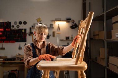 Orta yaşlı bir kadın marangozluk atölyesinde eski ahşap sandalyeyi süngerle zımparalıyor. mobilya yenileme, diy ve restorasyon konsepti