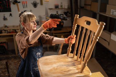 Kadın zanaatkar, evdeki atölyede elektrikli matkapla ahşap sandalyeyi monte ediyor. Boşluğu kopyala