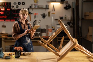 Atölyede çalışan kadın marangoz dijital tablet kullanarak mobilya ve ahşap ürünler yapıyor. Boşluğu kopyala