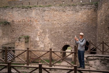Şapkalı yaşlı bir adam ortaçağ kalesini ziyaret ediyor. Modern akıllı telefon kullanarak fotoğraf çekiyor. Aktif emeklilik kavramı. Boşluğu kopyala